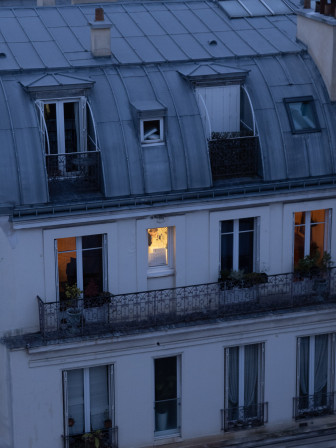 Une rangée de fenêtres allumées, deux en orangé sur les côtés, une plus jaune au centre. On aperçoit une femme en train de se brosser les dents.
